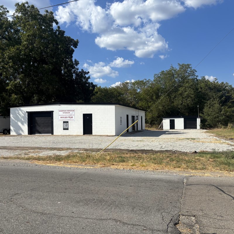 White storage building with a black roll-up door and a banner on a gravel lot, with a smaller building and trees behind it