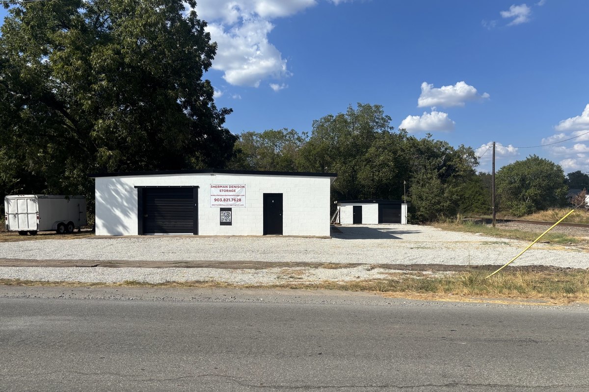 Straight-on view of the white storage building and a smaller one behind it on a gravel lot beside a street
