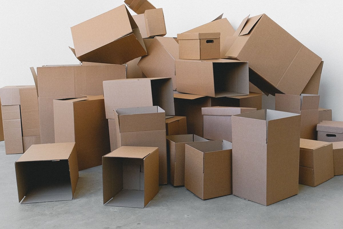 Heap of open and sealed brown cardboard boxes piled in the corner of a bare white room on a concrete floor