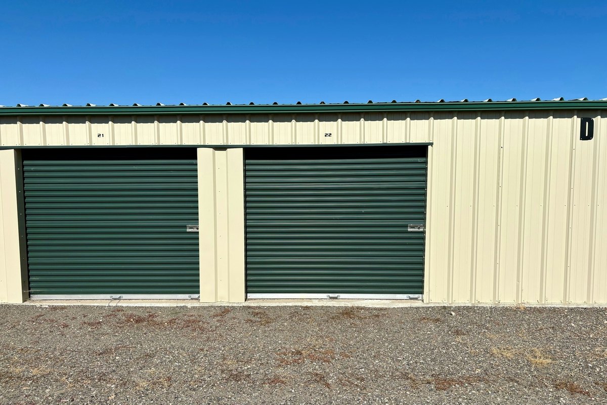 Cream metal storage building with two dark green roll-up doors on a gravel lot