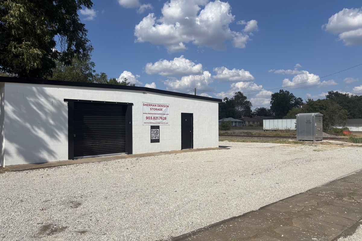 Long white cinder-block storage building with a black roll-up door and a steel door on a light gravel lot