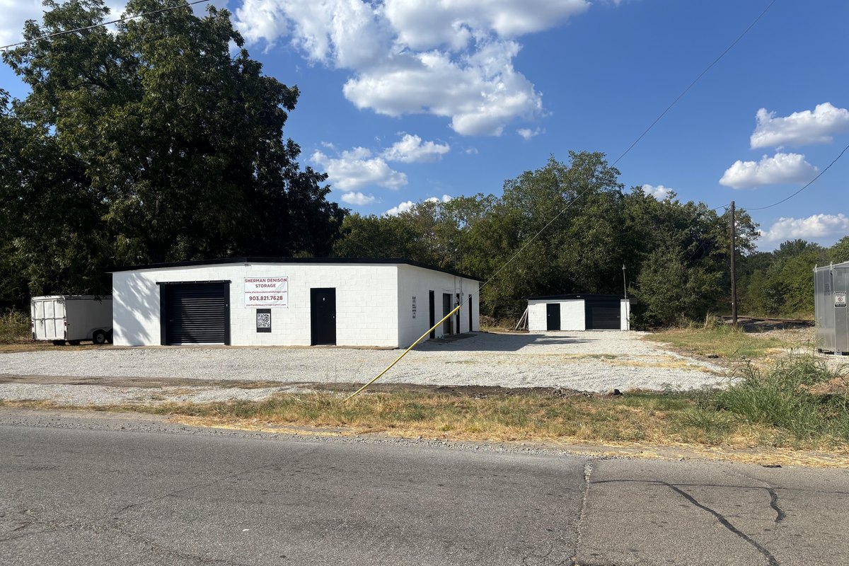 Angled view of the white storage building with its black roll-up door, a smaller building behind and gravel in front