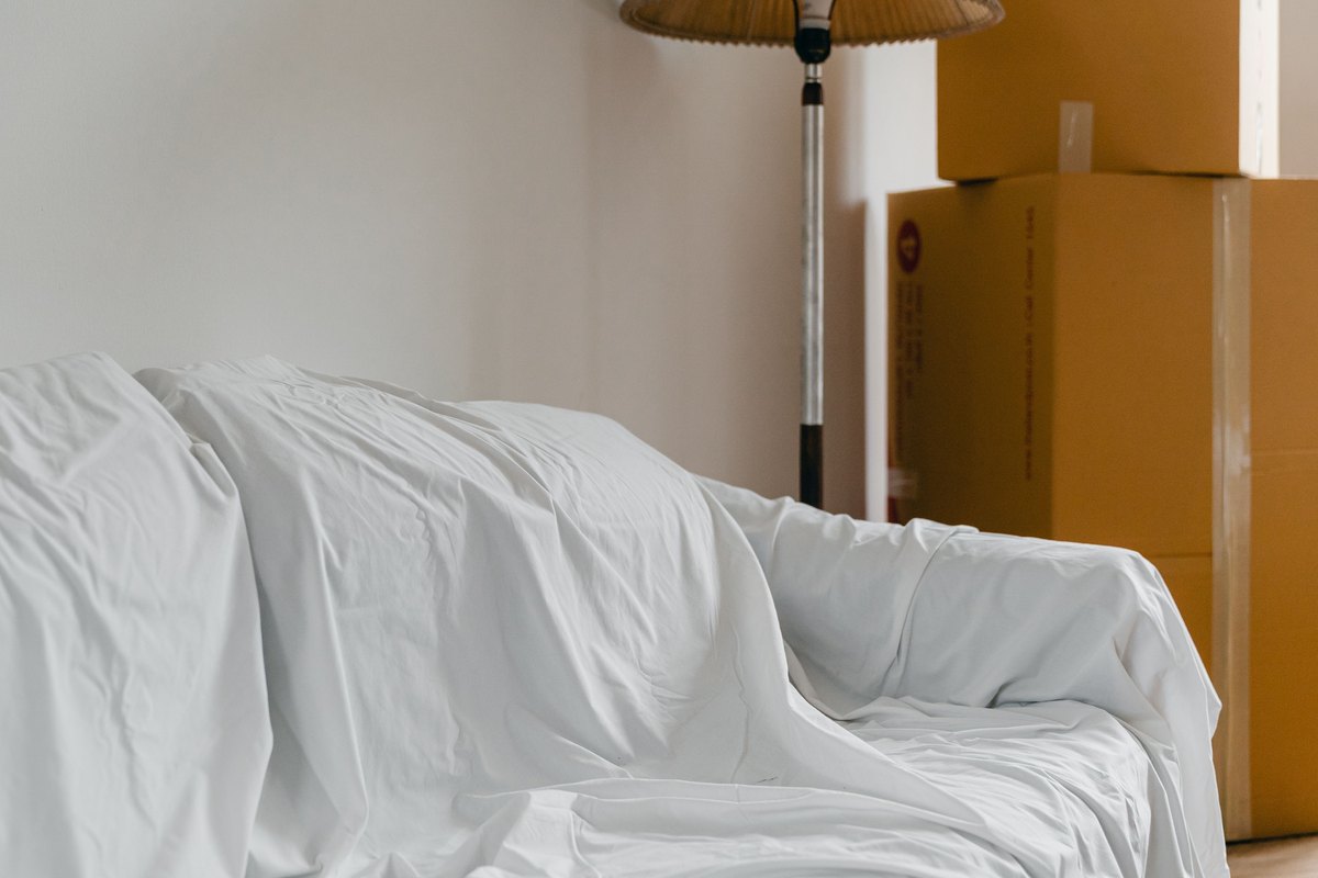 Sofa covered with a white sheet beside a floor lamp and stacked taped moving boxes in a bright room