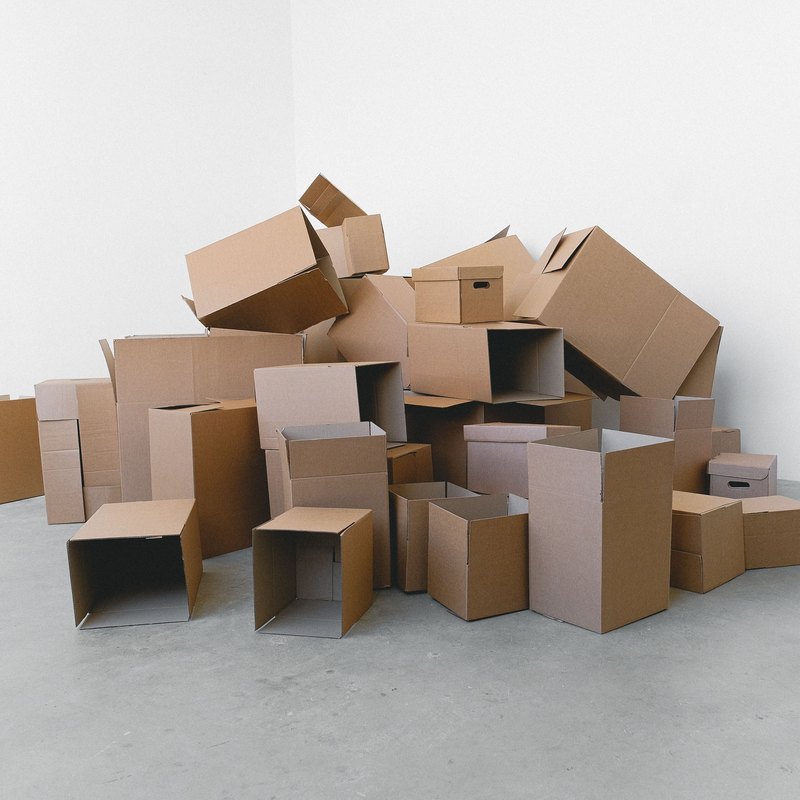 Heap of open and sealed brown cardboard boxes piled in the corner of a bare white room on a concrete floor