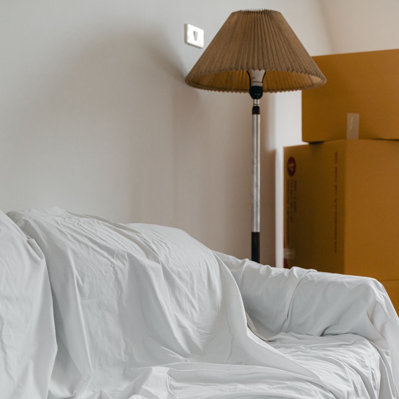 Sofa covered with a white sheet beside a floor lamp and stacked taped moving boxes in a bright room